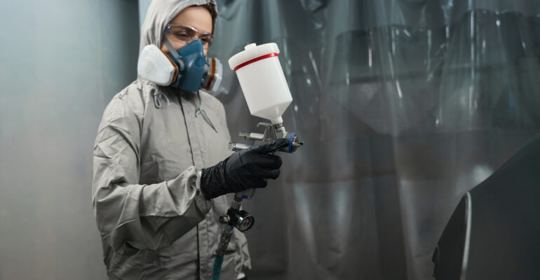 Photo of woman car technician holding spray gun indoors Professional female engineer in protective workwear lost in thought while spraying paint with airbrush on bodywork in service garage