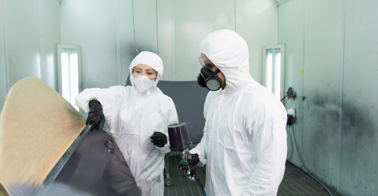 Workman in protective suit holding aerograph near colleague pointing with finger at car part in Workman in protective suit holding aerograph near colleague pointing with finger at car part in